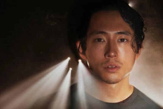 Steven Yeun stands against a wall with light coming through bullet holes