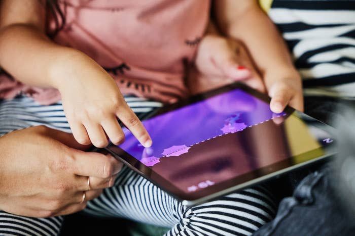 Child's hand touches tablet screen showing a game; adult hand holds tablet. Child is wearing striped pants