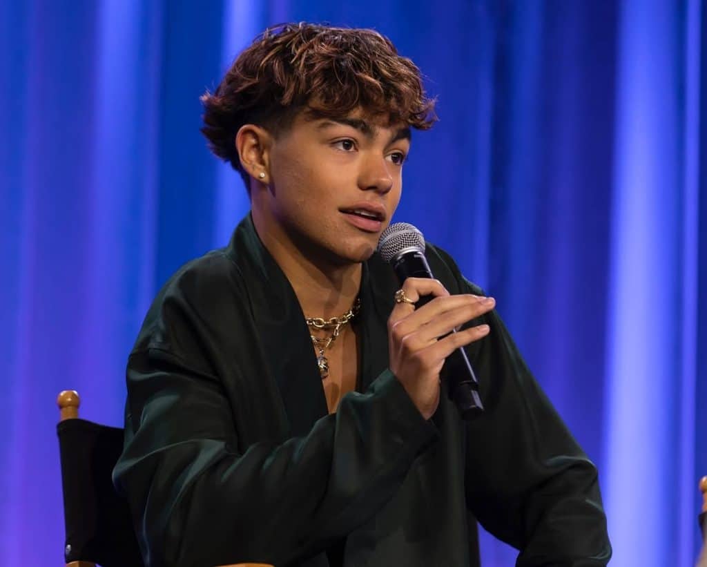 A person with short, curly brown hair holds a microphone while seated, wearing a dark green jacket and gold necklaces, in front of a blue and purple curtain backdrop at the Descendants Zombies Worlds Collide Tour. Free tickets may be available.