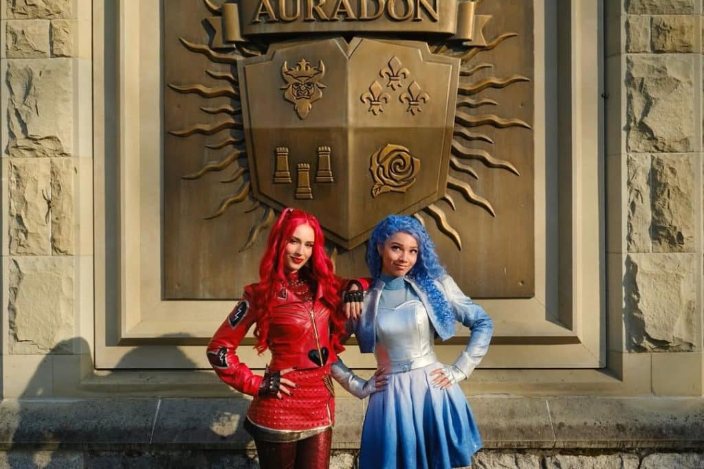 Two women in colorful costumes, one in red with long red hair and one in blue with long blue hair, smile before a large stone crest engraved with Auradon, celebrating the magic of Descendants at the Worlds Collide Tour.