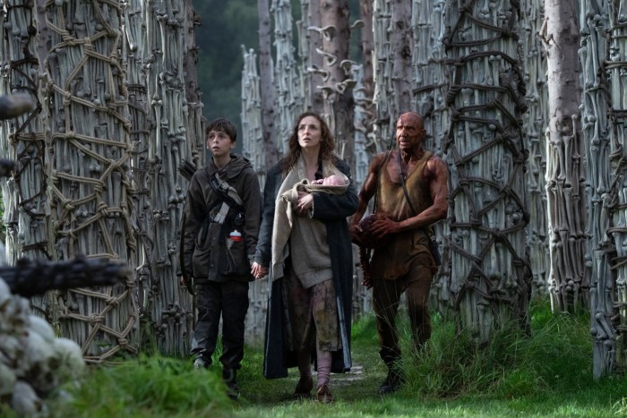 A boy in combat gear, a woman holding a baby, and a blood-splattered bald man in a vest walk through a forest, where the tree trunks have been covered in ropes and climbing pegs
